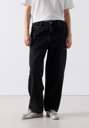 Oseba, oblečena v široke črne kavbojke, belo majico in srebrne superge, stoji ob enobarvnem svetlem ozadju.