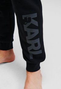 Fekete, puha textúrájú melegítőnadrág nagyméretű szürke logóval, amely az oldalon ott áll, hogy "KARL", a bokánál szűkített fazonnal.