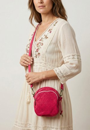 Kvinde iført cremefarvet kjole med broderede blomster og blonde detaljer, der holder en lille lyserød flettet crossbody-taske med rem.