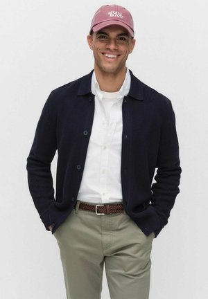 Marine gebreide cardigan over een wit shirt, beige chinos en een roze baseball cap met de tekst "SUN DAY". Met knopen en een gevlochten riem.