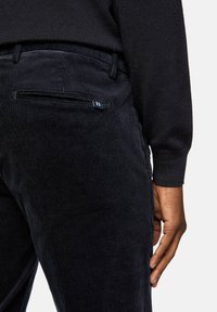 Dark corduroy pants with a ribbed texture, featuring a back pocket and a small blue logo tag. The material appears soft and durable.