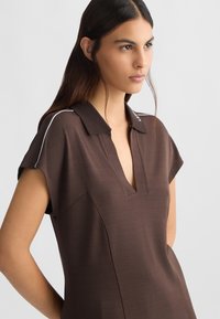 Abito polo marrone con maniche corte, scollatura a V e dettagli in piping bianco lungo le spalle. Tessuto liscio e vestibilità su misura.