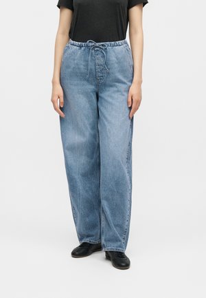 Άτομο που φοράει φαρδιά ανοιχτό μπλε τζιν με ελαστική μέση και κορδόνι, μαύρα παπούτσια και μαύρο κοντομάνικο τοπ.