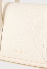 Crèmekleurige leren handtas met een getextureerd oppervlak. Voorzien van gouden merkopdruk "COCCINELLE" aan de voorkant en naaddetails langs de randen.