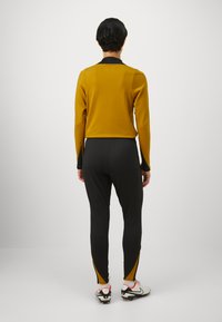 Sport antrekk med en sennepsgul langermet topp og svarte joggebukser. Joggebuksene har gule sidestriper. Brukt med hvite treningstøvler.
