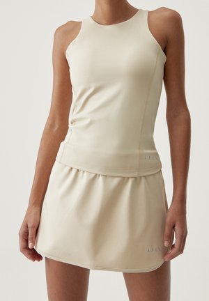 Vrouw draagt een beige, mouwloze, nauwsluitende top en bijpassende rok, staand met ontspannen handen langs haar zij tegen een lichte achtergrond.