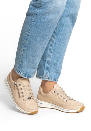 Sneakers in pelle beige con lacci e cerniera laterale indossate con jeans di denim azzurro chiaro su caviglie nude.