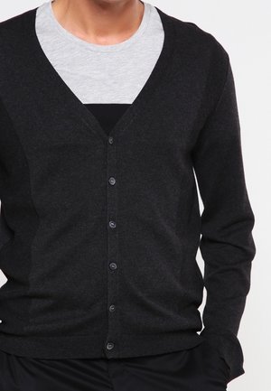 Homme portant un cardigan boutonné gris foncé sur un t-shirt gris clair à col rond, debout les mains dans les poches.