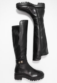 Bottes en cuir noir montantes jusqu'au genou, à texture lisse, semelle en caoutchouc épaisse, fermeture éclair latérale et accents dorés à la cheville.