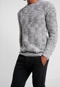 Gestrickter Pullover in Grautönen mit einem strukturierten Muster, Rundhalsausschnitt und gerippten Bündchen. Kombiniert mit schwarzen Hosen.