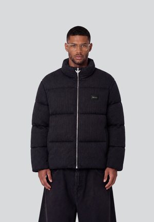 Mann mit klaren Brillen und einer schwarzen gesteppte Jacke mit Frontreißverschluss und kleinem Logo auf der Brust, steht vor einem einfarbigen Hintergrund.