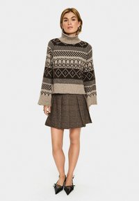 Beige- und braun gemusterter Rollkragenpullover mit geometrischen Designs, kombiniert mit einem braunen Faltenrock. Das Modell trägt schwarze spitze Absätze.