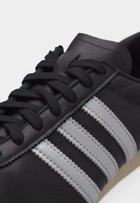 Μαύρο δερμάτινο αθλητικό παπούτσι με μαύρα κορδόνια και τρεις ανάγλυφες λευκές ρίγες κατά μήκος της πλευράς σε ανοιχτό γκρι φόντο.