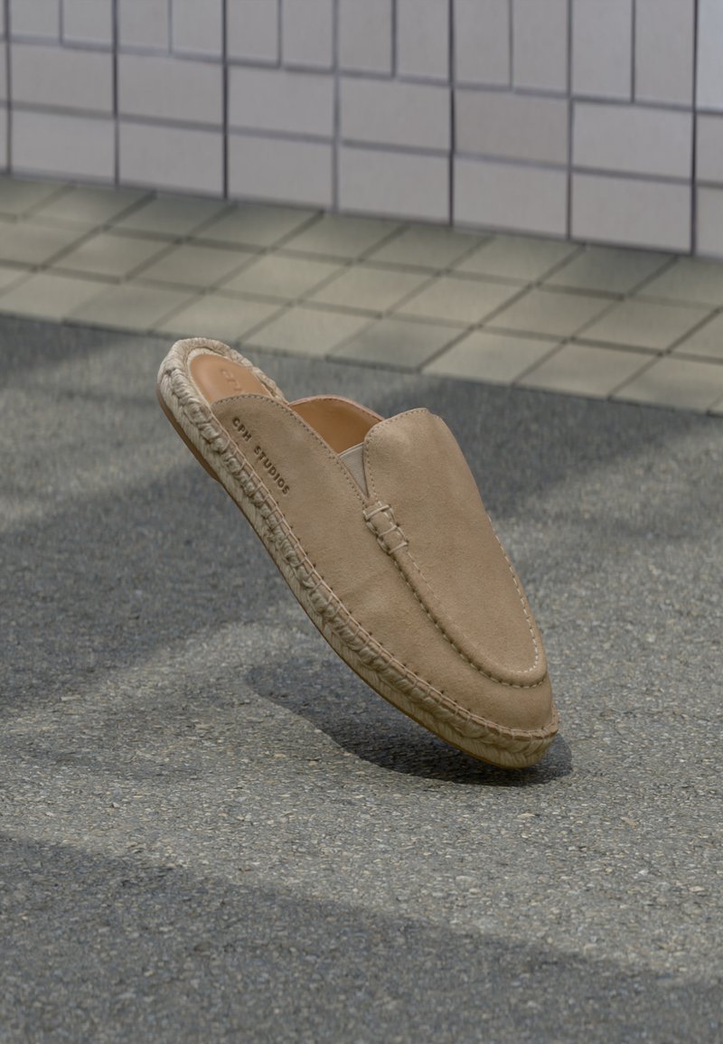 Chaussure à enfiler en daim marron clair avec semelle tressée flottant au-dessus de l'asphalte avec un mur carrelé en arrière-plan.