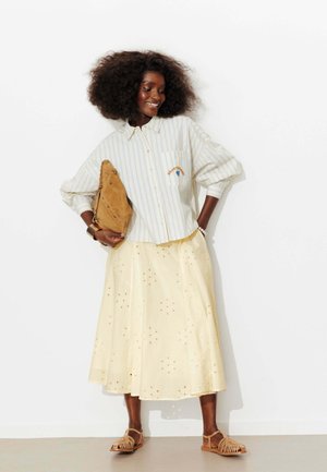 Femme aux cheveux bouclés et volumineux portant une chemise rayée boutonnée, une jupe midi jaune à œillets, des sandales marron clair et tenant une pochette marron.