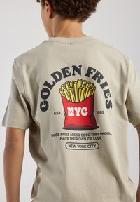Beige short-sleeve t-shirt featuring a graphic of a red container of fries with "GOLDEN FRIES NYC" text and slogan on the back.