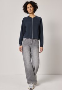 Femme aux cheveux bouclés portant une veste zippée bleu marine, un jean gris délavé à jambes larges et des baskets blanches, debout devant un fond uni.