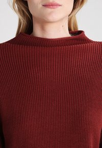 Bordeauxrode geribbelde trui met een hoge, afgeronde halslijn. De textuur is fijngebreid en heeft een glad, rekbaar materiaal.