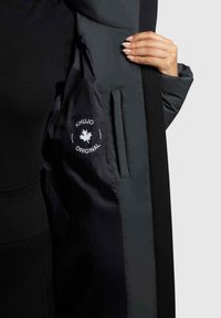 Czarna kurtka zewnętrzna z gładką, ciemną podszewką, na której znajduje się białe logo. Widoczna kieszeń i dopasowany krój.
