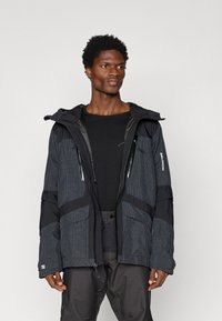 Bărbat îmbrăcat cu o jachetă cu model închis și glugă, peste o cămașă neagră și pantaloni închiși la culoare, stând în fața unui fundal deschis.