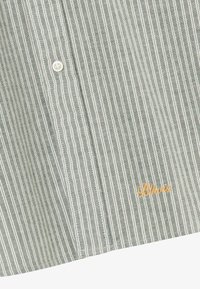 Grünes und weißes vertikal gestreiftes Hemd mit Knopfleiste, weißen Knöpfen und kleinem gelbem gesticktem "Bikkio"-Logo in der Nähe des Saums.