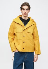 Gul lett jakke med hette, knappelukking foran og to brystlommer, båret over en svart t-skjorte og kombinert med blå jeans.