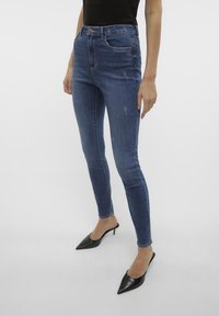 Tamno plave uske traperice od denima s visokom stražnjicom, s minimalnim oštećenjima, pet džepova i klasičnom kopčom na gumb.