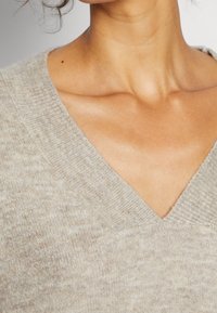 Jasnobrązowy, teksturowany sweter z dzianiny z dekoltem w serek. Materiał wydaje się miękki, posiada subtelny wzór melanżowy i prążkowane krawędzie.