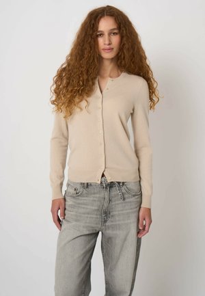 Kobieta o długich kręconych włosach, ubrana w beżowy kardigan z guzikami i jasnoszare, wysokim stanem jeansy, stojąca na jednolitym tle.