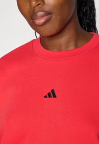 Rdeča bombažna majica s dolgimi rokavi s krožnim izrezom in črnim Adidasovim logotipom na prsih. Gladka tekstura brez vzorcev ali poudarkov.