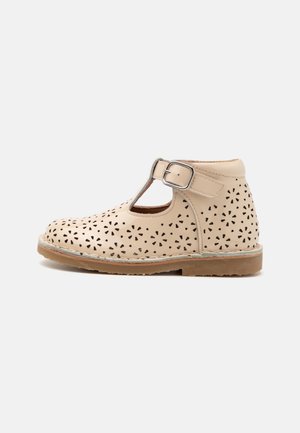 Zapato de cuero beige con patrones florales recortados, diseño de barra en T, hebilla metálica ajustable y suela de goma marrón.