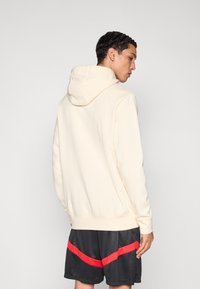 Světle béžová mikina s kapucí na šňůrku, dlouhými rukávy a žebrovanými manžetami, v kombinaci s černými šortkami s červeným diagonálním pruhem.