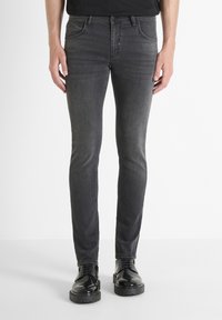 Uske slim-fit sivi traperice s blagom teksturom, pet džepova i zatvaranjem na gumbe, uparene s crnim sjajnim cipelama.