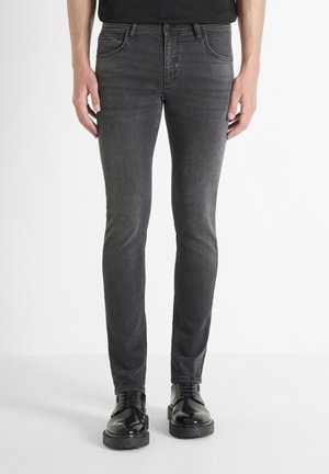 Uske slim-fit sivi traperice s blagom teksturom, pet džepova i zatvaranjem na gumbe, uparene s crnim sjajnim cipelama.