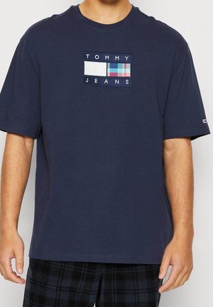 Homme portant un t-shirt bleu marine Tommy Jeans avec un patch logo rectangulaire sur la poitrine et un pantalon à carreaux noir et gris.