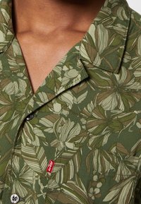 Chemise verte avec motif de feuilles tropicales marron et beige et étiquette rouge Levi's près des boutons, montrée sur la partie supérieure de la poitrine et du cou d'une personne.