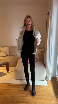 Lichtgrijze donsjas met witte accenten, zwart shirt en legging. Zwarte kniehoge laarzen. Houten vloer, gestreepte beige bank op de achtergrond.