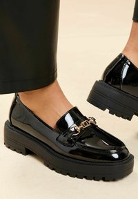 Fekete lakkbőr mokaszinok vastag fekete talppal, aranylánc díszítéssel a pánton és texturált gumitalppal.