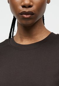 Κοντινό πλάνο ενός ατόμου με σκούρα πλεξούδες που φοράει μαύρο μπλουζάκι με στρογγυλή λαιμόκοψη, με φόντο απλό.