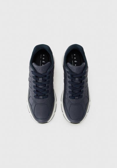 Un paio di sneakers blu scuro con lacci neri e suole bianche, viste dall'alto su uno sfondo grigio chiaro.