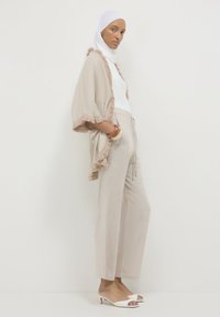 Donna che indossa un hijab bianco, uno scialle beige con frange, una maglietta bianca, pantaloni beige a gamba larga e sandali con tacco bianchi, in piedi su uno sfondo bianco uniforme.