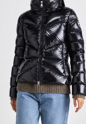 Doudoune noire brillante avec fermeture éclair à l'avant, portée sur un pull en maille marron et un jean bleu clair sur une personne debout.