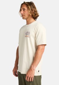 T-shirt en coton beige clair à manches courtes, featuring un imprimé graphique en violet et orange sur la poitrine. Coupe décontractée avec un col ras du cou.