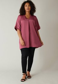 Tunika color prugna con scollatura a V, vestibilità ampia e maniche corte arrotolate, abbinata a pantaloni neri aderenti e sandali con tacco a blocco neri.