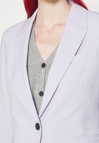 Personne portant un blazer violet clair par-dessus un cardigan gris en tricot boutonné, avec des cheveux roux visibles derrière le col.