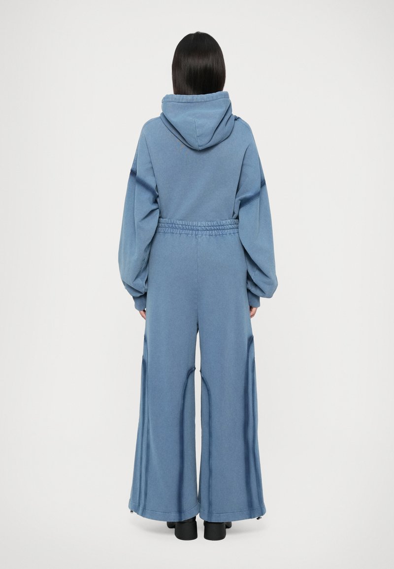 Kvinna som står med ryggen mot kameran, iklädd en lös blå huvtröja och vida byxor tillsammans med svarta klackskor på vit bakgrund.