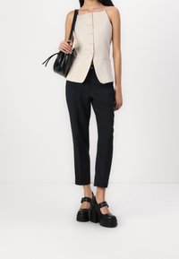 Chaleco entallado beige con botones, combinado con pantalones negros ajustados. La modelo lleva zapatos negros de tacón grueso y sostiene un pequeño bolso negro.