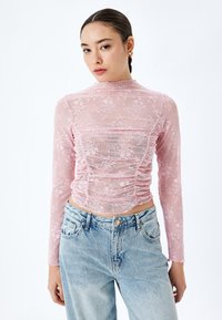 Rosa durchsichtige Langarmbluse mit floralem Spitzenmuster, Raffung an der Vorderseite und hohem Hals, kombiniert mit hellblauen High-Waist-Jeans.