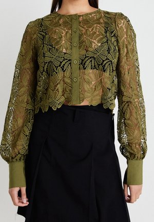 Długi, koronkowy crop top w oliwkowej zieleni z kwiatowym wzorem i guzikami z przodu, noszony na czarnym crop topie i zestawiony z czarną spódnicą.