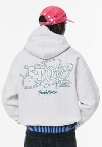 Felpa grigia con una grande stampa grafica sul retro, che presenta testo e disegni in teal. Orlo a coste blu a righe visibile nella parte inferiore.